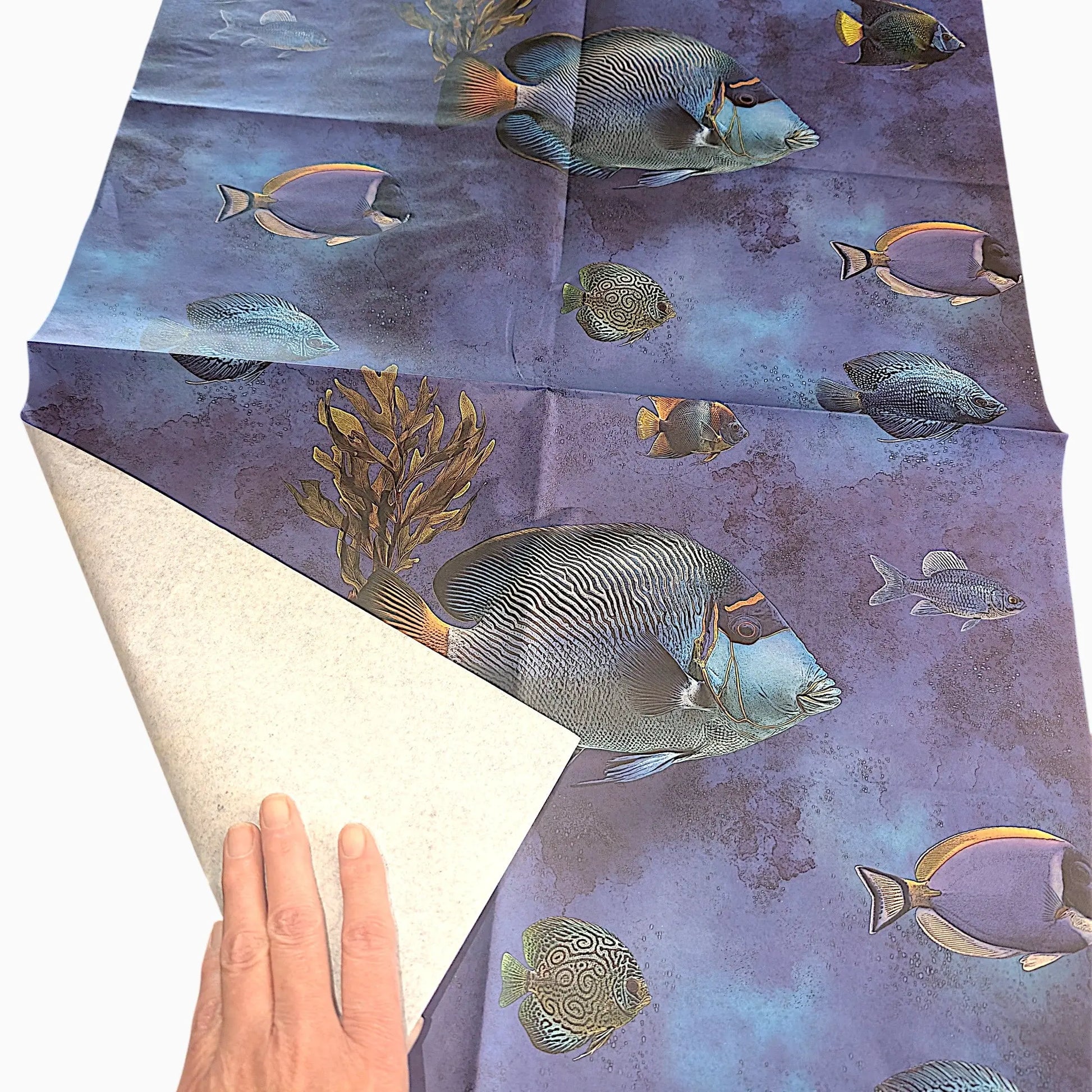 A square fabric or paper sheet with an underwater fish pattern, shown with a hand lifting one corner to reveal the off-white reverse side. The print features blue, purple, and teal tropical fish on a mottled ocean background.