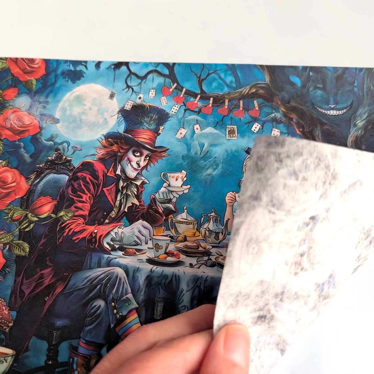 A hand holds a translucent, sheet of rice paper in front of a colorful printed illustration featuring the Mad Hatter from Alice in Wonderland at a tea party under a full moon.