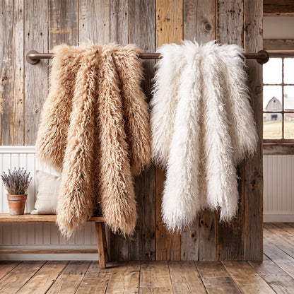 Two fluffy faux-shearling style throws hanging from a rustic metal rod against a wood plank wall, with one throw in warm beige and the other in off-white. A wooden bench with pillows and a small potted plant sits below.
