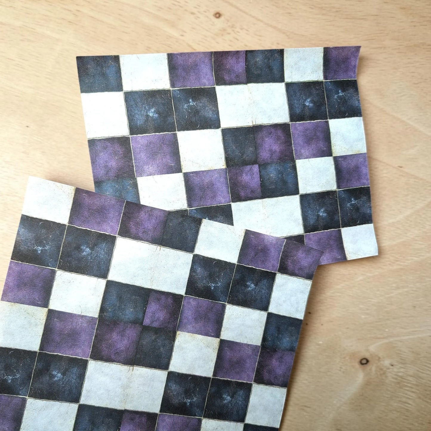 Two square checkered patterned cards or tiles with a purple, black, and off-white checkerboard design, displayed on a light wood surface.
