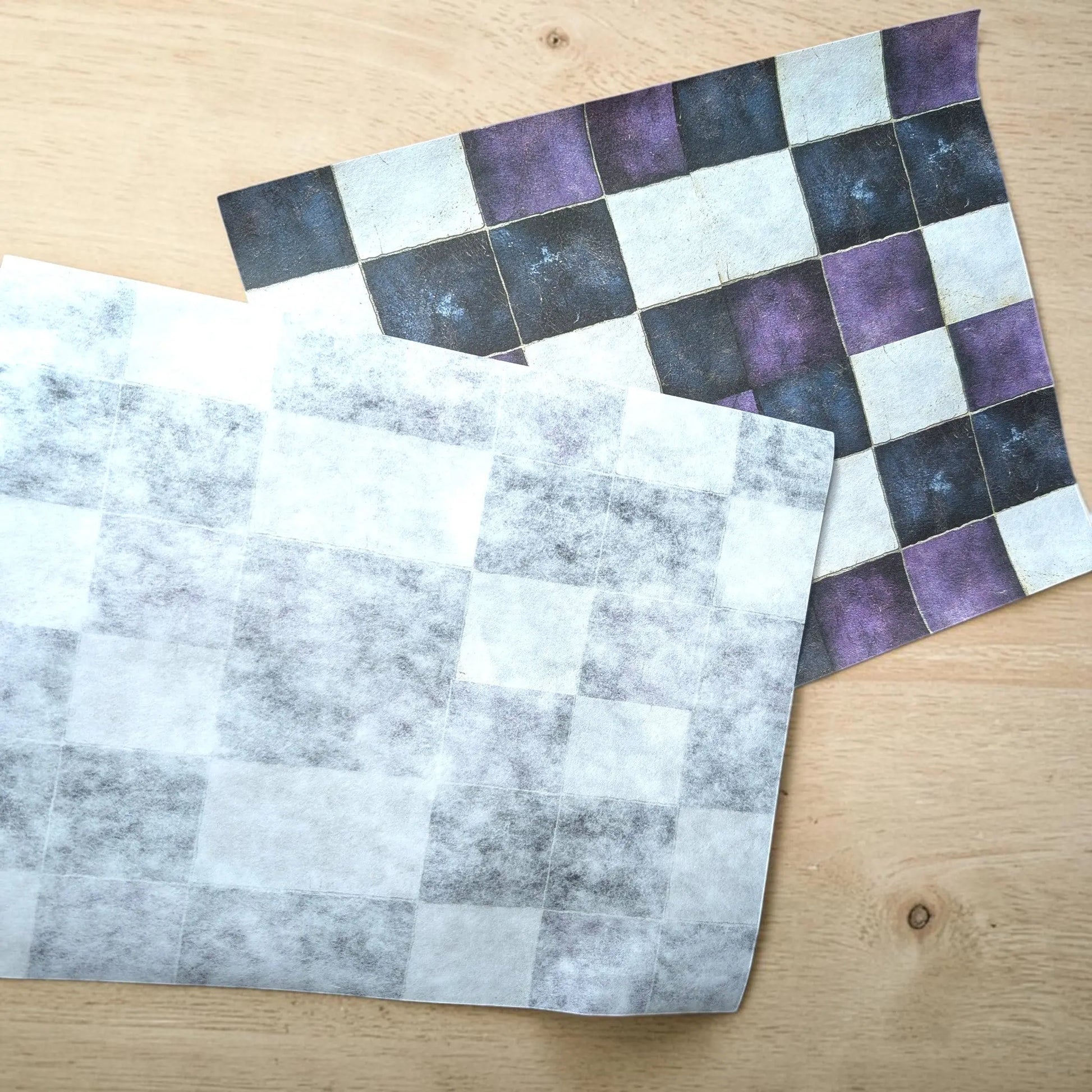 Two square checkered fabric placemats with a checkerboard pattern, one in gray and white and the other in purple, black, and white, displayed on a light wood tabletop.
