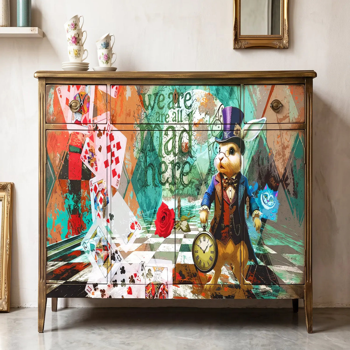 A vintage-style wooden sideboard or dresser with a highly detailed, colorful Mad Tea Party illustration on the front doors, featuring a rabbit in a top hat, playing cards, a rose, and the text 'we are all mad here'. The cabinet has a distressed gold/bronze frame and is styled in a room setting with teacups on top and a framed mirror on the wall.