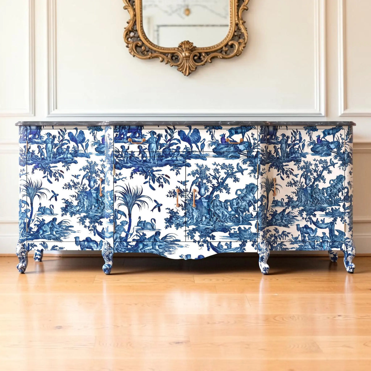 A three-front sideboard or credenza covered in a classic blue and white toile pattern, set against a white paneled wall with a gold-framed mirror above and a light wood floor below.