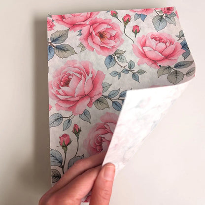 A hand holds a corner of a tissue paper sheet of floral wrapping paper with pink roses and green leaves on a light background, showing the white adhesive backing.