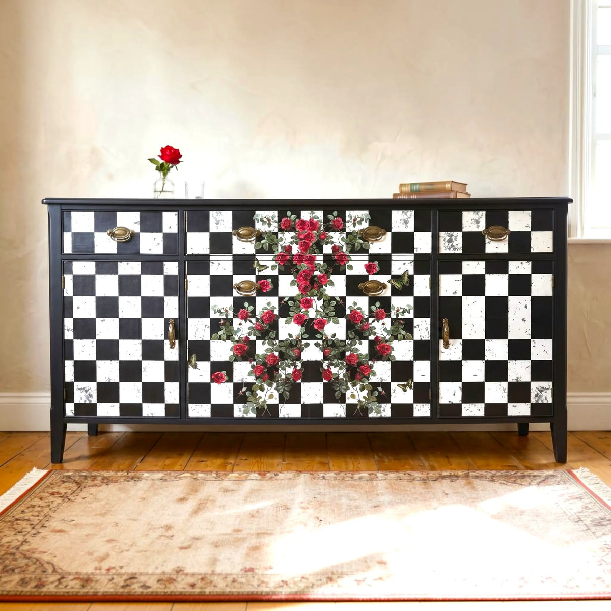 A black and white checkered sideboard cabinet with red rose floral motifs in the center, styled in a warm room with a rug, wooden floor, and a vase of roses on top.